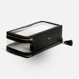 Anya Hindmarch Logo In-Flight Case: a rectangular black toiletry bag with clear panels, gold-tone zippers, and a small gold bow emblem, featuring a secure zip closure.