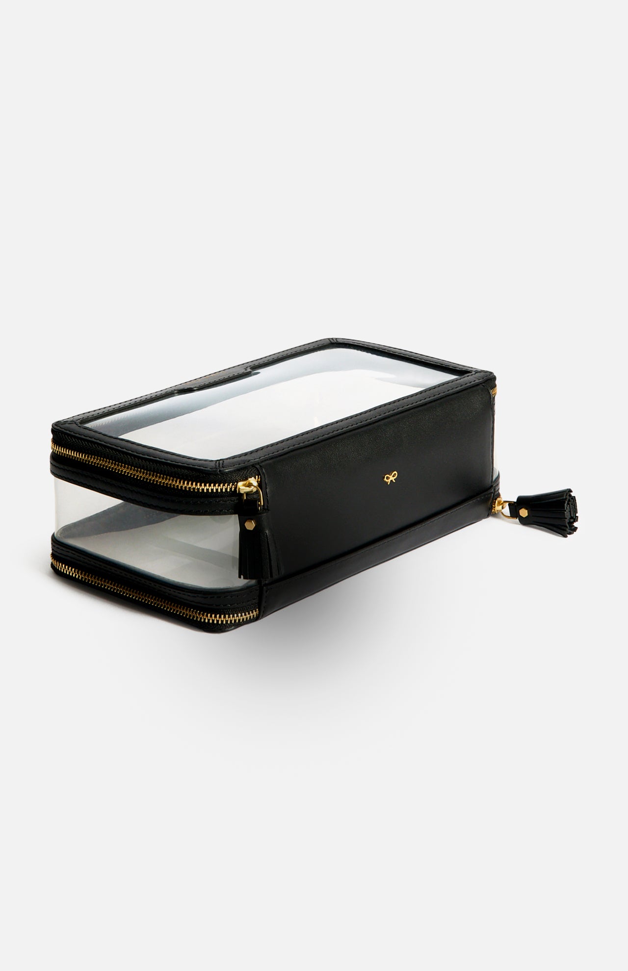 Anya Hindmarch Logo In-Flight Case: a rectangular black toiletry bag with clear panels, gold-tone zippers, and a small gold bow emblem, featuring a secure zip closure.