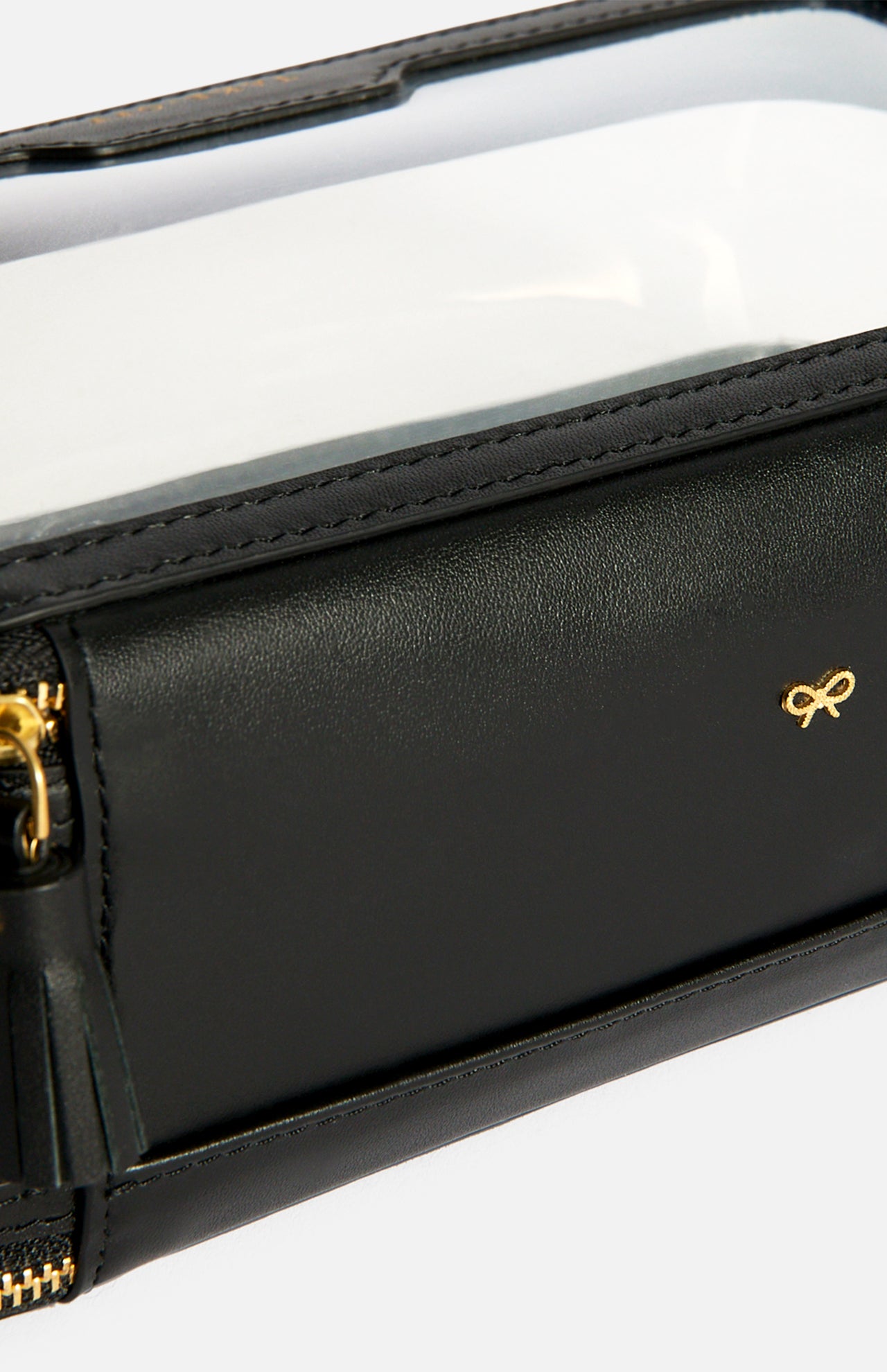 Anya Hindmarch Logo In-Flight Case in black with a zippered closure, clear plastic top, and small gold bow emblem.
