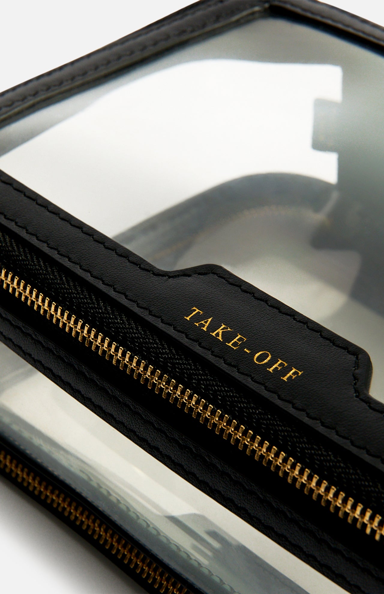 Clear, rectangular Anya Hindmarch Logo In-Flight Case with black leather trim, a gold-tone zip closure, and the word TAKE-OFF embossed in gold on the front.