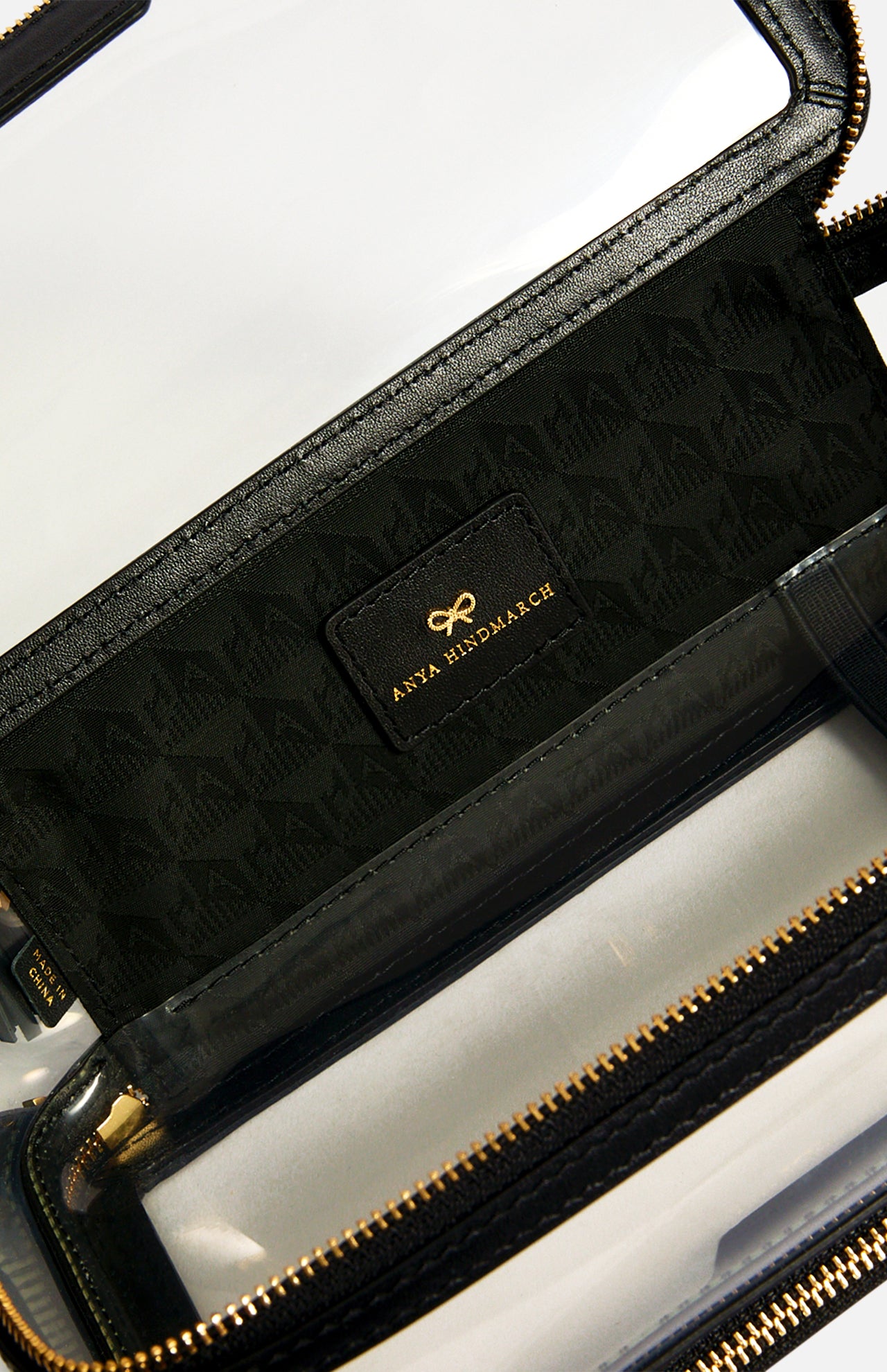 Anya Hindmarch Logo In-Flight Case in a clear design with black trim, gold-tone hardware, and a black interior lining featuring a bow logo and gold text.