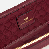 Close-up of the burgundy Logo In-Flight Case interior, showing a rectangular leather patch embossed with a gold bow logo and ANYA HINDMARCH text against a patterned lining.