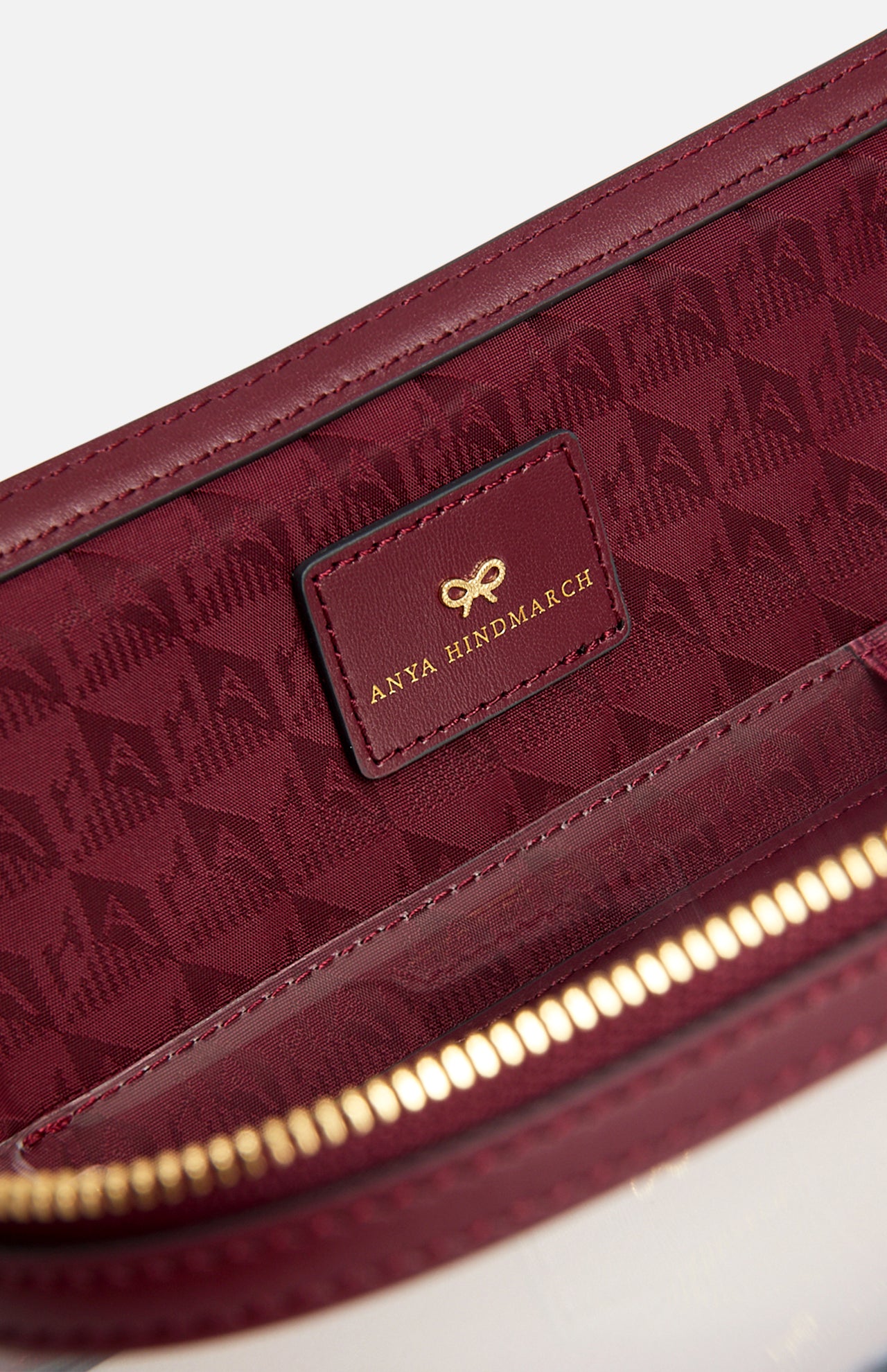 Close-up of the burgundy Logo In-Flight Case interior, showing a rectangular leather patch embossed with a gold bow logo and ANYA HINDMARCH text against a patterned lining.