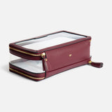 Anya Hindmarch Logo In-Flight Case: a rectangular, burgundy cosmetic case made in Italy featuring clear panels, gold zippers, and a small gold bow emblem on the front.