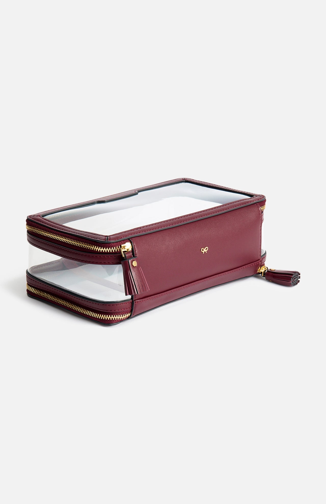 Anya Hindmarch Logo In-Flight Case: a rectangular, burgundy cosmetic case made in Italy featuring clear panels, gold zippers, and a small gold bow emblem on the front.
