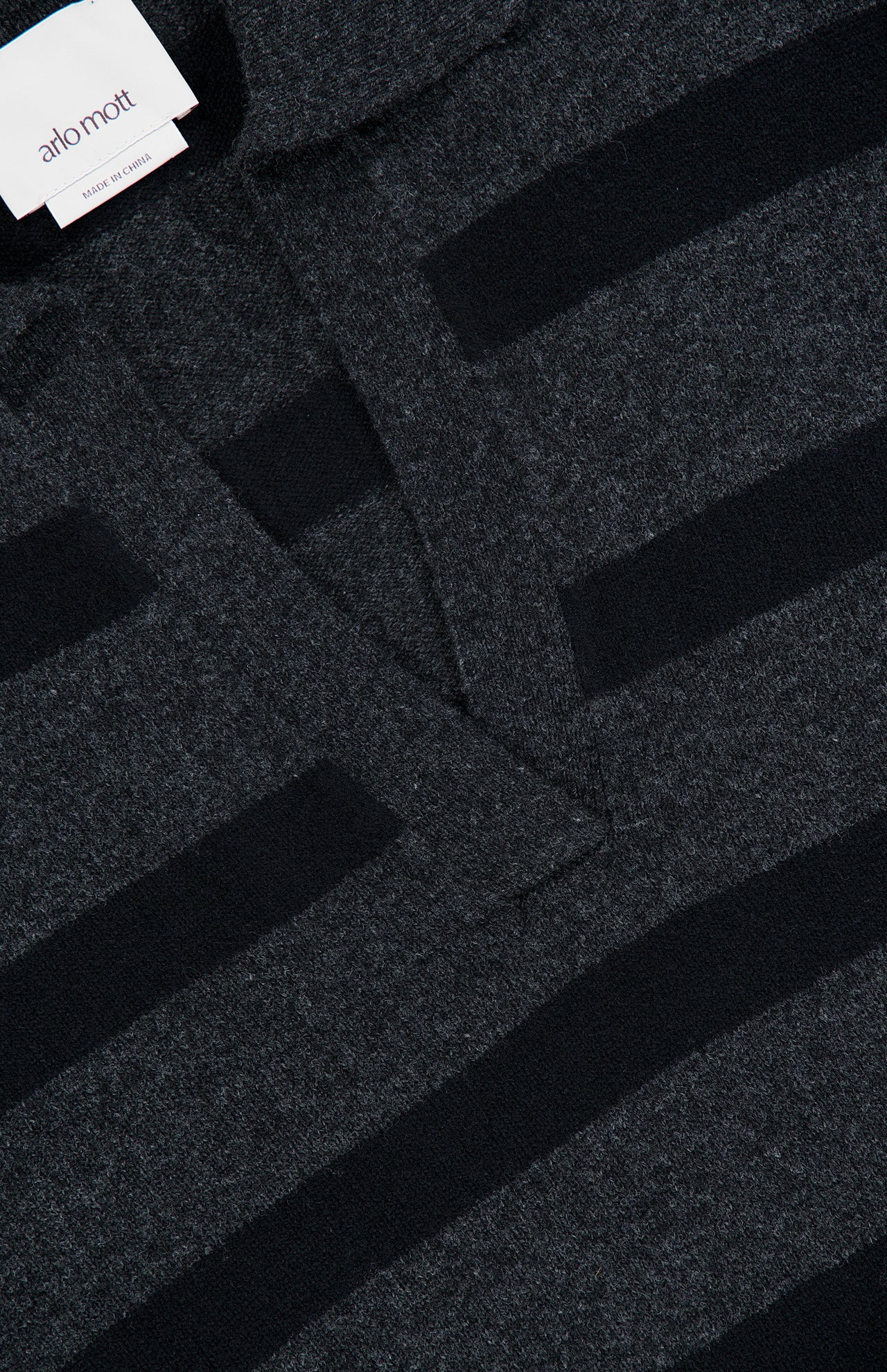 Close-up of the Allen Wool Cashmere Oversized Sweater in dark grey and black charcoal stripes, featuring a collar and v-shaped neckline. A visible white tag on the collar displays the brand name ARLO MOTT.