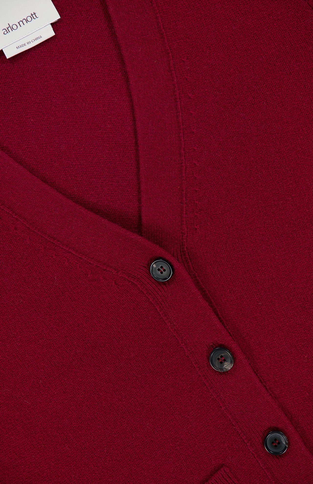 Close-up of a deep red v-neck Bleeker Shaped Cashmere Cardigan by ARLO MOTT, featuring black buttons and a visible collar label with the brand’s name. The knit cashmere fabric and classic design highlight the luxurious look, making it an elegant Italian yarn cardigan.