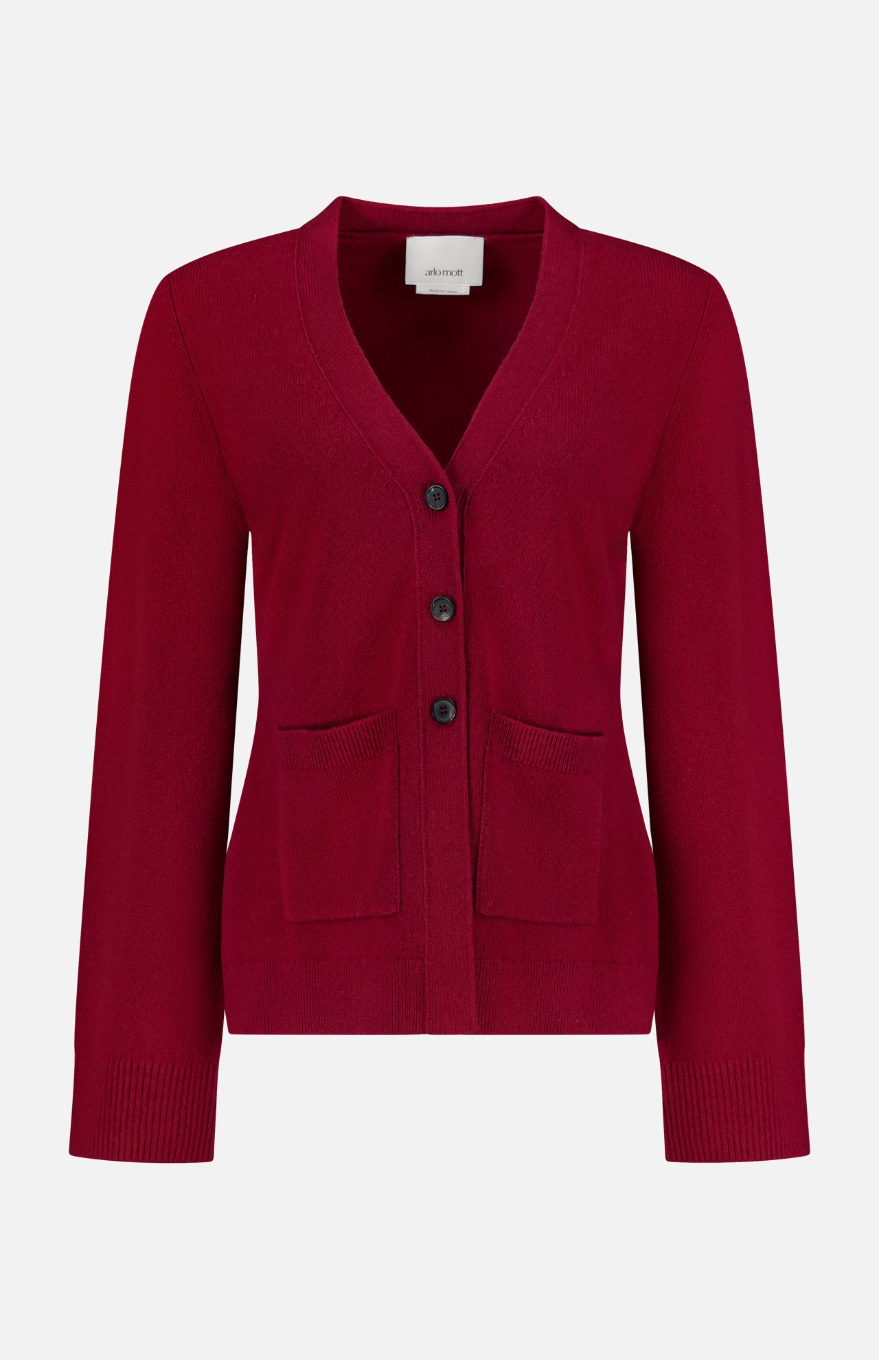 The Bleeker Shaped Cashmere Cardigan by ARLO MOTT is a red V-neck design, expertly knitted from luxurious Italian cashmere. It features long sleeves, three black buttons down the front, and two front pockets. Ribbed detailing at the cuffs and hem adds a refined touch. Presented against a plain white background.