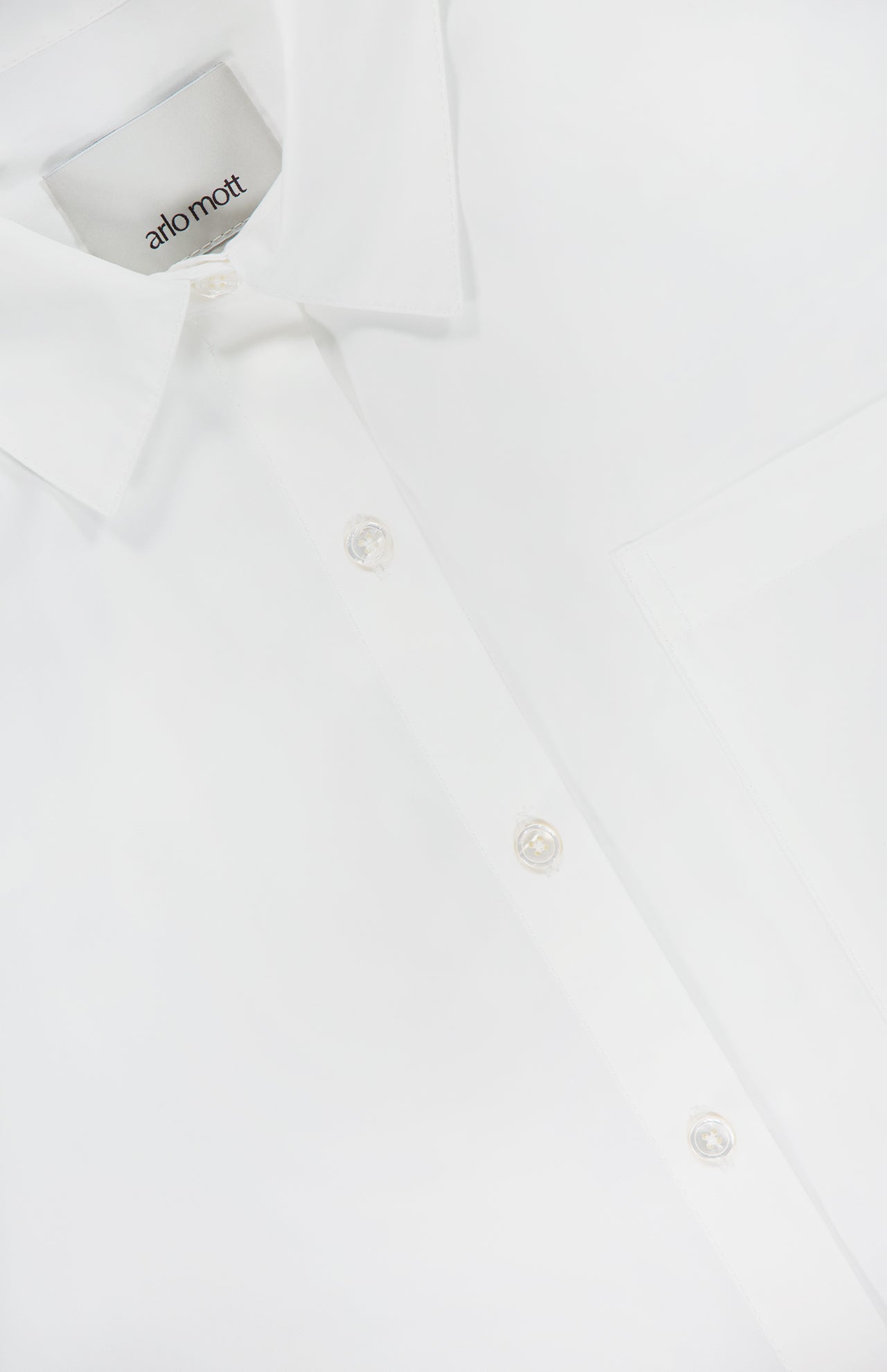 Close-up of the Clark Split Back Top by ARLO MOTT, a crisp white poplin shirt featuring a pointed collar, Mother of Pearl buttons, and a visible front pocket. The inside label shows the ARLO MOTT brand.