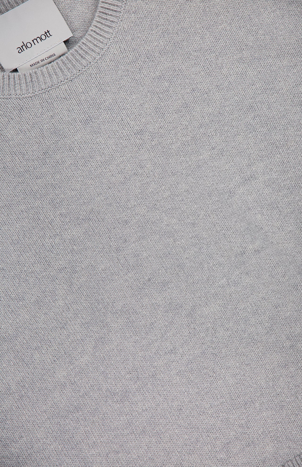 A close-up of the Clarkson Short Sleeve Crewneck Shirt by ARLO MOTT, a minimalist light grey knit with a ribbed neckline. A white clothing label reading ARLO MOTT is sewn into the collar of this Italian wool blend top.