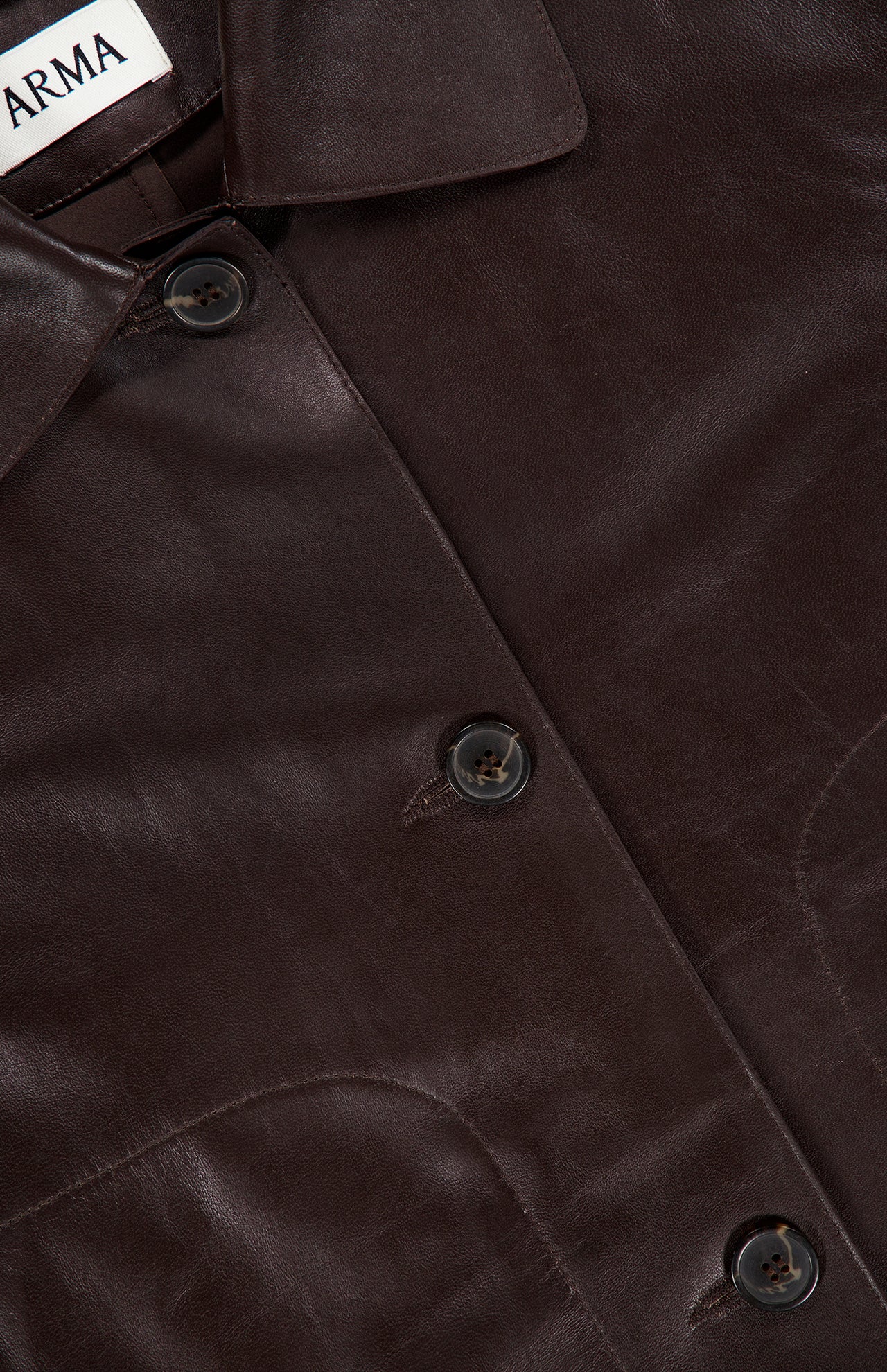 Close-up of the ARMA Emy Lamb Butter Coat, a dark brown jacket crafted from soft lamb leather, featuring three black buttons and a partially visible white ARMA label at the collar, highlighting its tailored silhouette.
