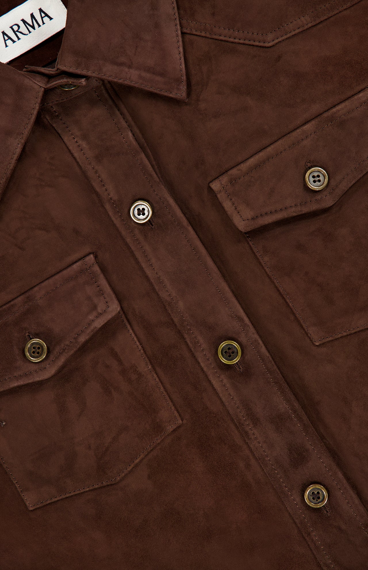 Close-up of the ARMA Faye Goat Suede Sturdy Coat, a brown suede button-up shirt-style coat featuring two chest pockets and visible buttons. A partial clothing label with the text ARMA is visible in the upper left corner.