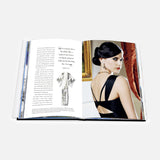 An open copy of ASSOULINE’s “Dressing For The Dark” displays text and a sketch on the left page, while on the right page, a woman in a black dress with an elegant updo and earrings is seated and glancing over her shoulder—evoking iconic fashion moments curated by celebrity stylist Kate Young.