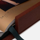 A close-up of ASSOULINE’s "Football: The Impossible Collection," an open, brown leather-bound book with a textured cover, dark green ribbon bookmark, and a page showing text and football history alongside a historical painting.