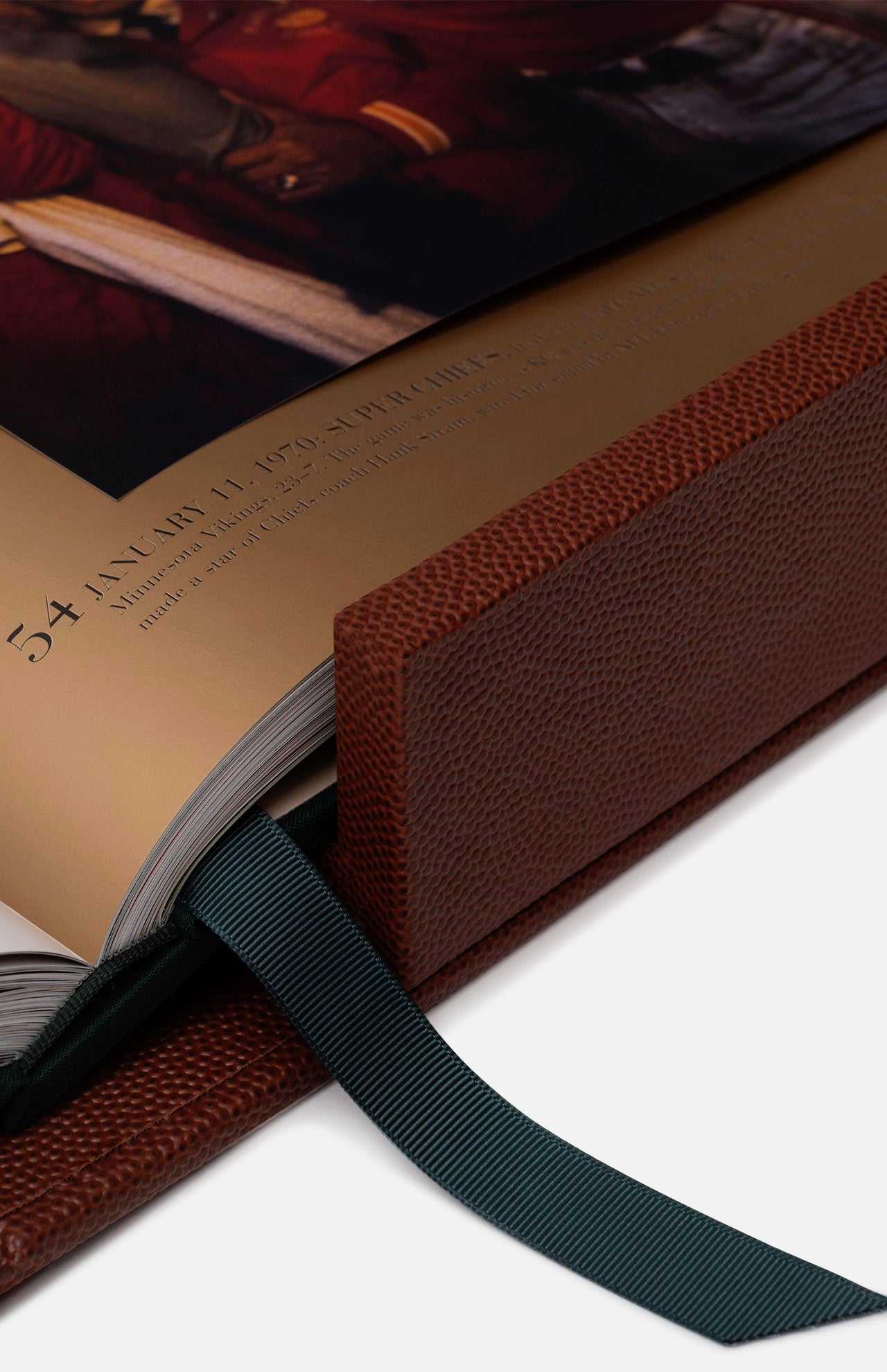 A close-up of ASSOULINE’s "Football: The Impossible Collection," an open, brown leather-bound book with a textured cover, dark green ribbon bookmark, and a page showing text and football history alongside a historical painting.
