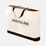 A large beige tote bag with black handles, featuring ASSOULINE printed boldly on the front. With a dark brown base and a white item inside, it's ideal for carrying the "Football: The Impossible Collection" book by ASSOULINE.