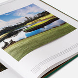 An open copy of ASSOULINE’s "Golf: The Impossible Collection" displays a beautiful golf course photograph with water, green fairways, trees reflected in the pond, and a blue sky overhead. Text is visible beneath the image.
