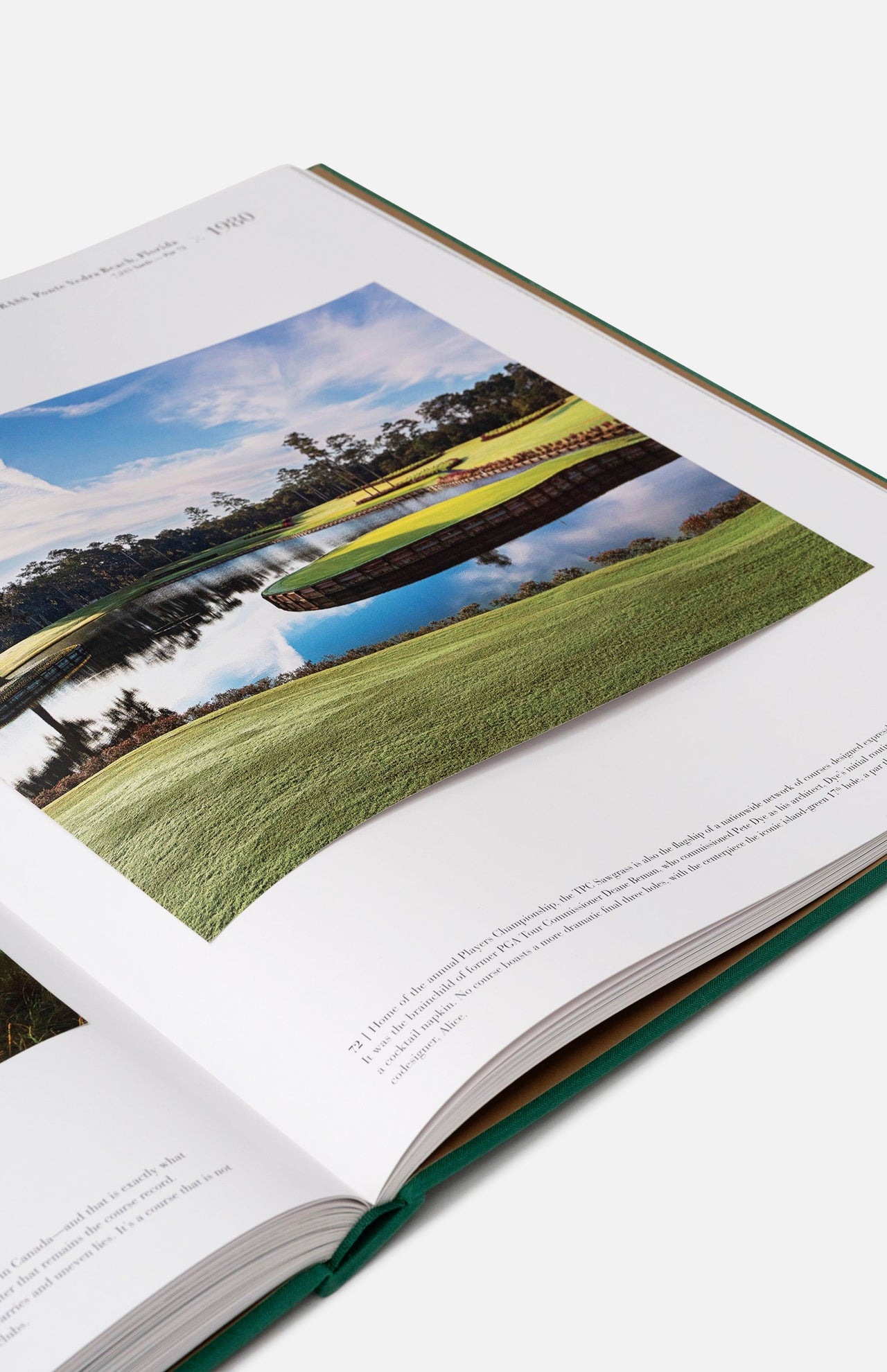 An open copy of ASSOULINE’s "Golf: The Impossible Collection" displays a beautiful golf course photograph with water, green fairways, trees reflected in the pond, and a blue sky overhead. Text is visible beneath the image.