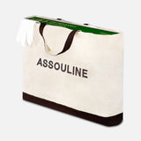 A large beige tote bag with black handles and ASSOULINE branding holds Golf: The Impossible Collection by ASSOULINE and a pair of white gloves peeking out.