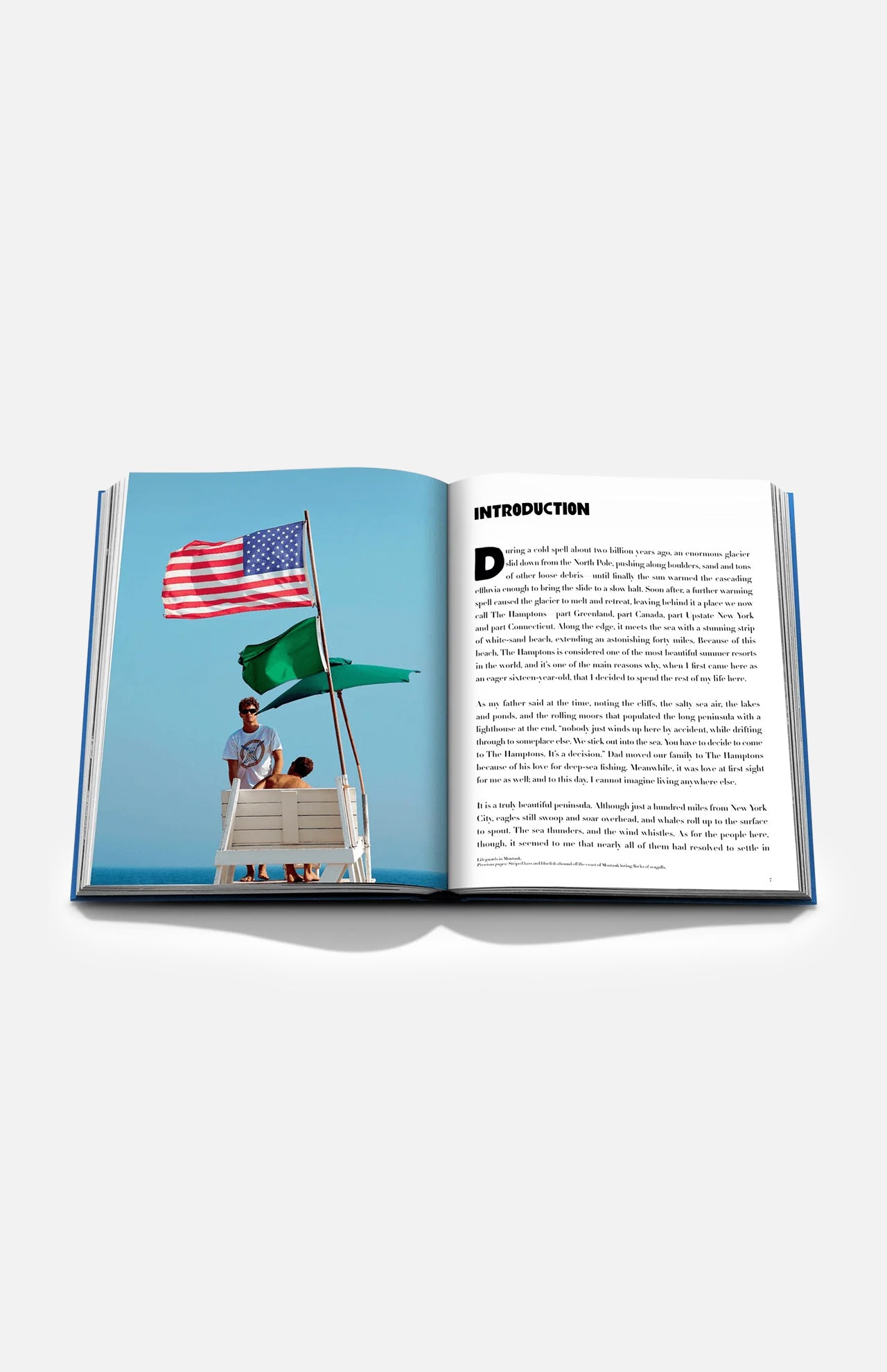 An open book, Hamptons Private by ASSOULINE, displays a photograph of two people on a lifeguard stand with American and green flags, embodying the luxury lifestyle of the Hamptons. Opposite the image, the right-hand page features introductory text—making this volume an ideal addition to any coffee table book collection.