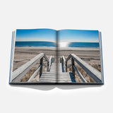 An open copy of ASSOULINE’s “Hamptons Private” displays a photograph of a wooden staircase descending to a sandy beach, with the ocean and blue sky beyond—perfectly capturing the quintessential Hamptons lifestyle.