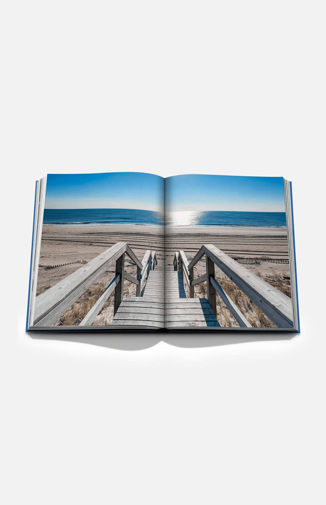 An open copy of ASSOULINE’s “Hamptons Private” displays a photograph of a wooden staircase descending to a sandy beach, with the ocean and blue sky beyond—perfectly capturing the quintessential Hamptons lifestyle.