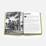 A book titled *Miami Beach* by ASSOULINE lies open on a white background, its striking yellow-edged pages showcasing a black-and-white photograph of people beneath palm trees on the left, and an Introduction section on the right. The imagery hints at art deco architecture and the vibrant cultural scene of Art Basel Miami Beach, capturing the city’s allure.