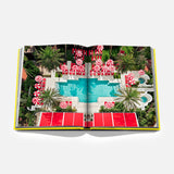 An open copy of “Miami Beach” by ASSOULINE rests on a white background, displaying a vivid aerial image of a pool encircled by palm trees and red-and-white umbrellas—capturing the vibrant allure of Miami travel and reflecting the city’s iconic Art Deco architecture.