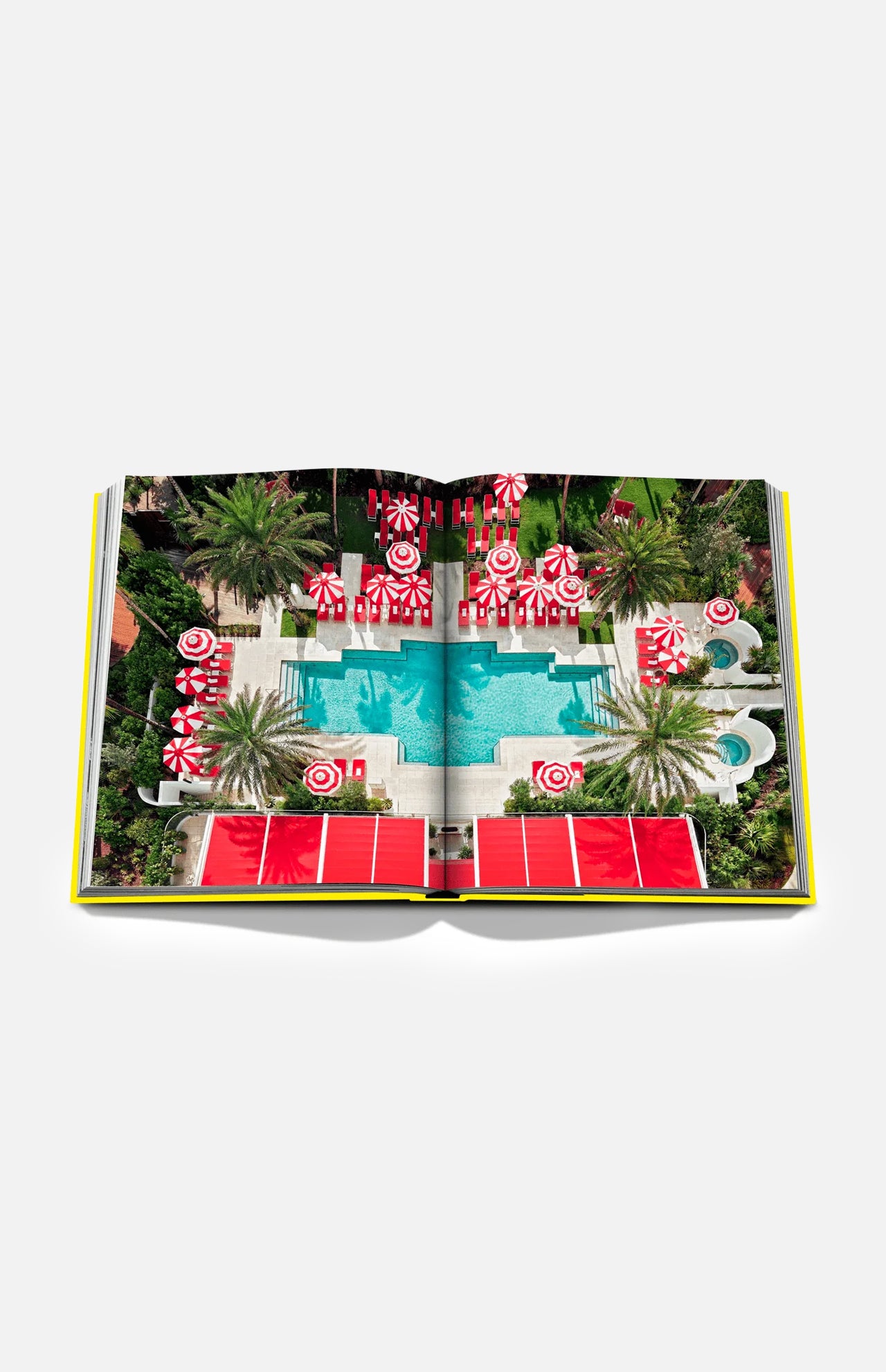 An open copy of “Miami Beach” by ASSOULINE rests on a white background, displaying a vivid aerial image of a pool encircled by palm trees and red-and-white umbrellas—capturing the vibrant allure of Miami travel and reflecting the city’s iconic Art Deco architecture.