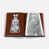 An open copy of *The Art of Tequila: Spirit of Mexico Book* by Assouline shows silver decorative objects—an ornate bottle with a face on the left page and an abstract, geometric Mexican-inspired design on the right.