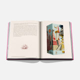 The Art of Tequila: Spirit of Mexico Book by Assouline rests on a white surface, opened to show text with a red plant illustration on the left and a vibrant, Mexican-inspired abstract artwork with plants and birds on the right.
