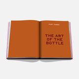 An open book titled "The Art of Tequila: Spirit of Mexico" by Assouline displays bold, dark text on orange pages, highlighting collectible tequila bottles and their relation to Mexican art.