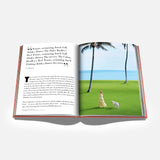 An open book from ASSOULINE titled “Palm Beach” displays text on the left page and, on the right, a photo of a woman in a long dress walking a white dog across green grass near palm trees and the ocean.