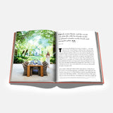 An open book from ASSOULINE titled “Palm Beach” displays a photograph of an outdoor dining table set amidst a lush garden, evoking the exclusive charm of Palm Beach. A woman stands nearby, while the opposite page features text with a prominent quote.