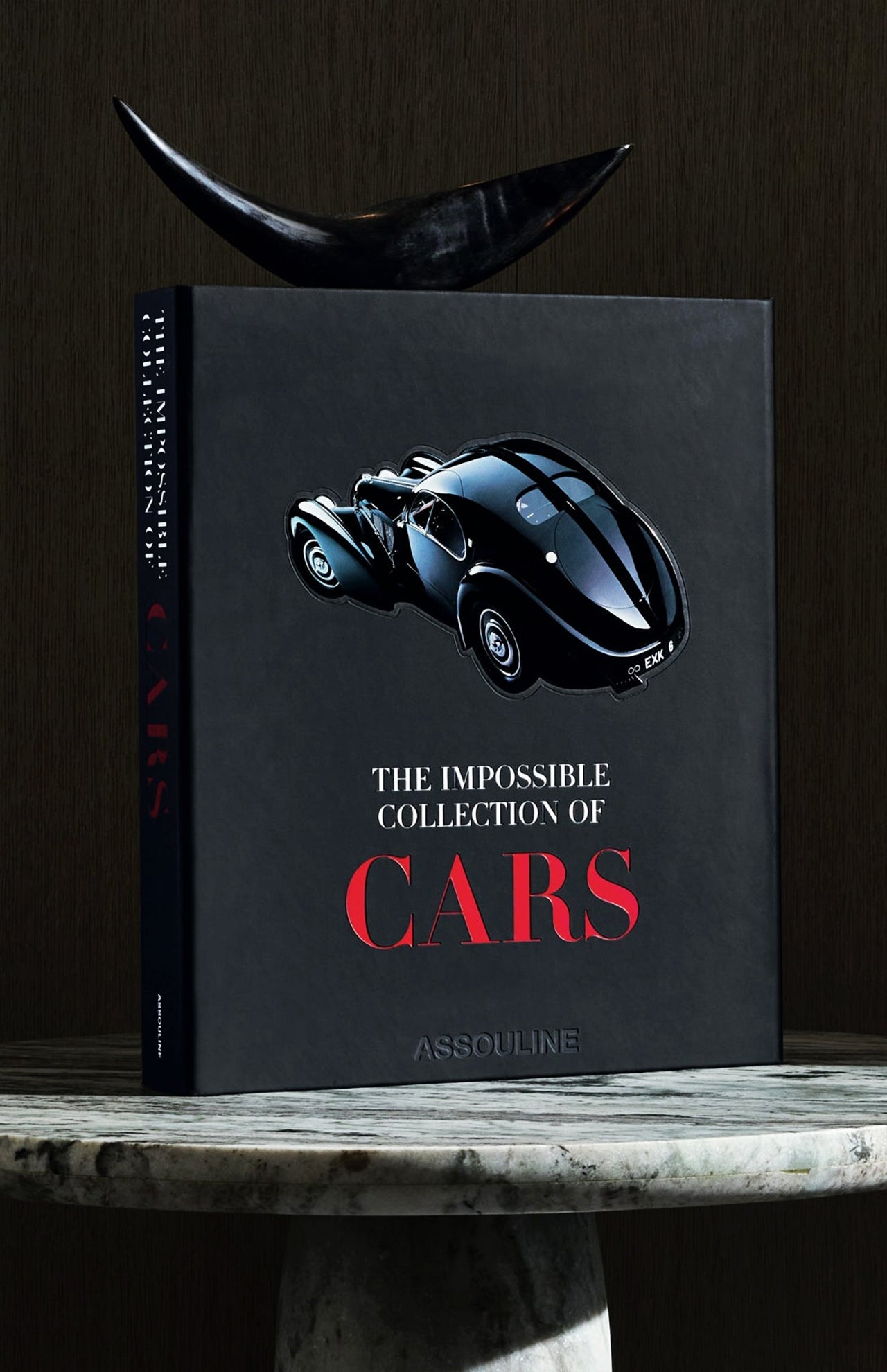 The ASSOULINE "The Impossible Collection of Cars" black coffee table book is displayed upright on a marble surface, topped with a decorative black sculpture.