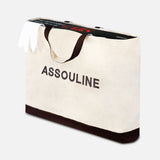A large white ASSOULINE canvas tote with black handles and trim holds a pair of white gloves and "The Impossible Collection of Cars" book by ASSOULINE, both partially visible inside the bag.