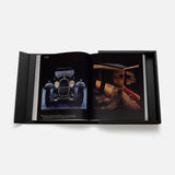 The Impossible Collection of Cars by ASSOULINE is a large hardcover book with a black cover and matching slipcase, open on a white surface to display vintage cars and luxurious interiors.