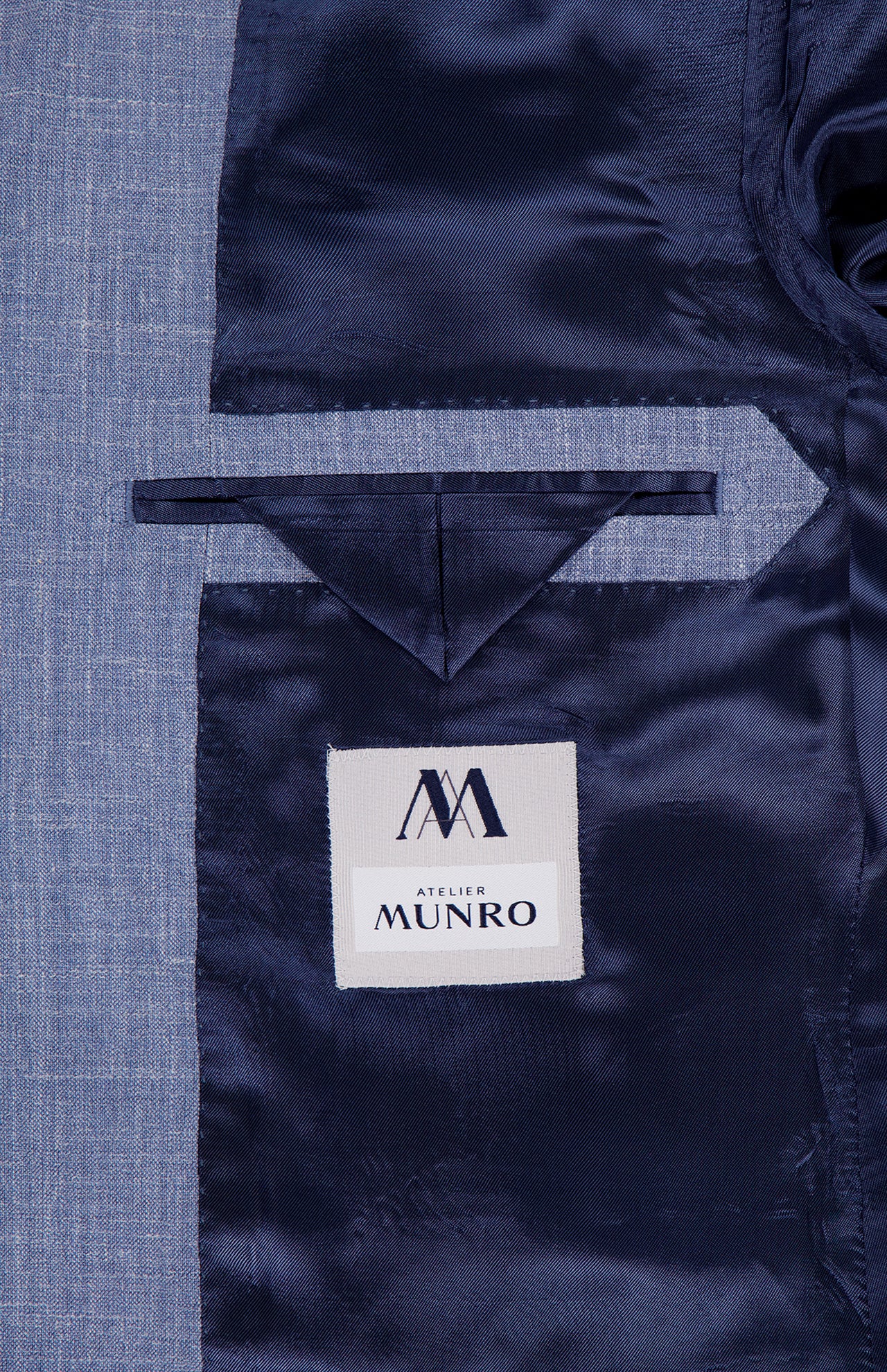 Close-up of the inside of the Melange Wool Silk and Linen Suit by ATELIER MUNRO, featuring a light blue lining, a pocket with a triangular flap, and a white label with the brand’s stylized AM logo.