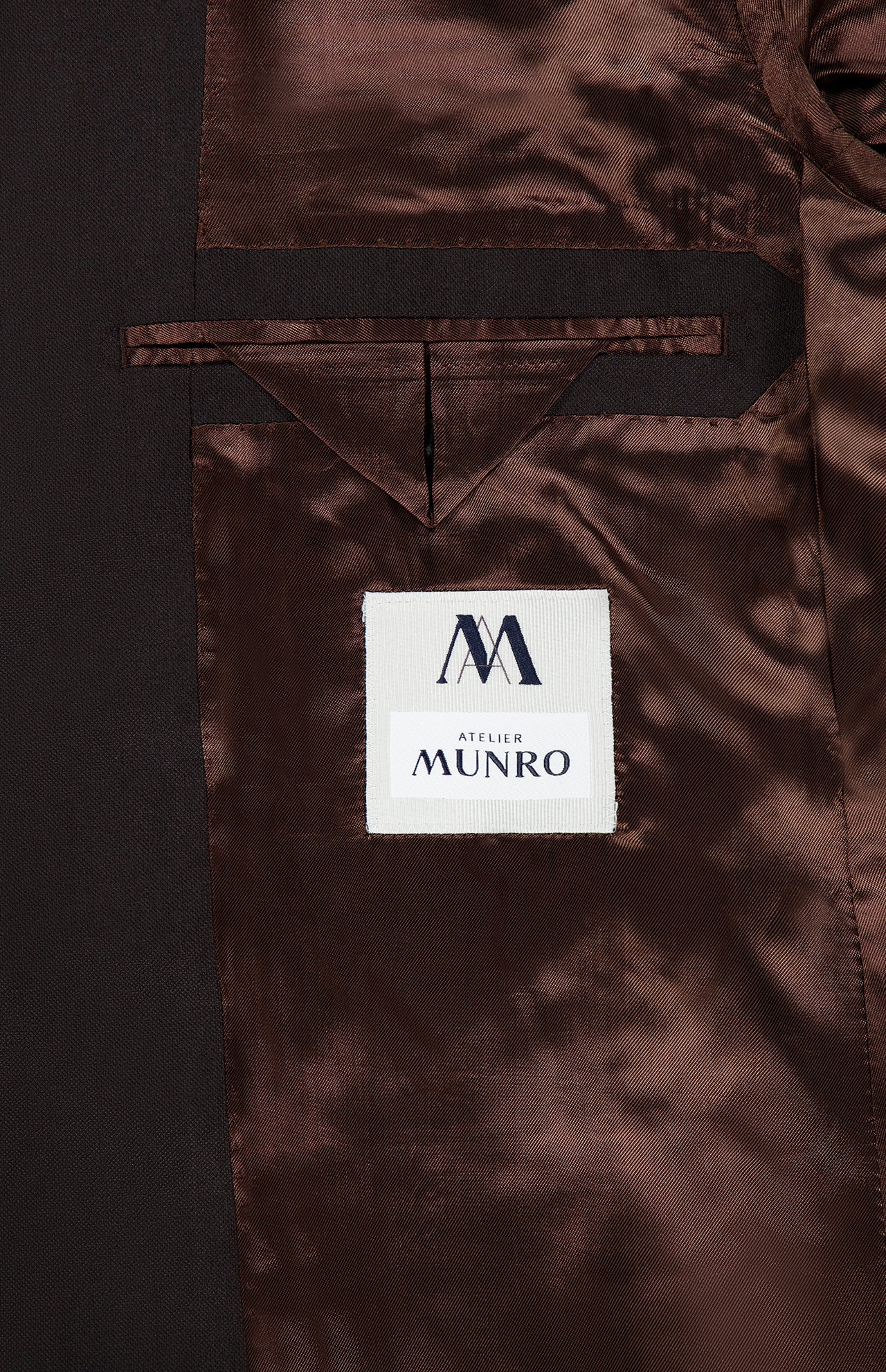 The inside of the Wide Notch Lapel Solid Suit by ATELIER MUNRO features shiny brown lining, a white Atelier Munro label, and a small chest pocket above it—a refined detail in this formal suit.
