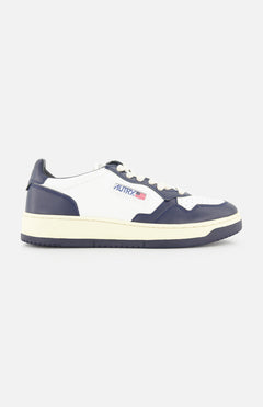 The AUTRY Medalist Low Man Leather Sneaker features white and navy panels, cream sole and laces, with the Autry logo and an American flag patch on the side—evoking classic 1980s tennis style. Displayed in profile against a white background.