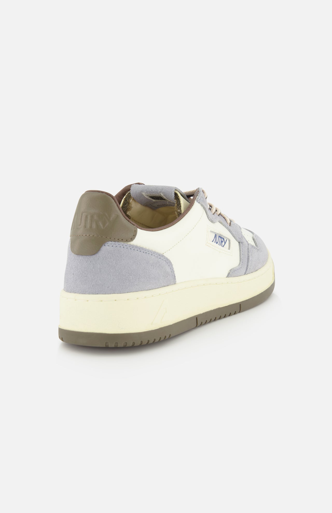 The AUTRY Medalist Low Man Leather Suede Sneaker is displayed from a rear side angle on a plain white background, showcasing its stylish combination of white, light gray, and beige hues. Inspired by classic 1980s tennis shoes, this sneaker features a suede heel detail, platform sole, rubber outsole, and a distinctive branded patch on the side.
