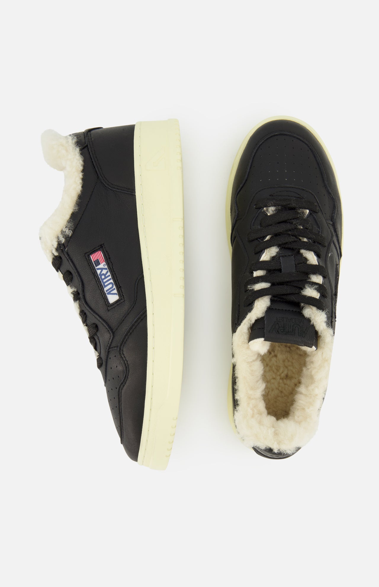 Two black Medalist Low Man Shearling Sneakers by AUTRY, featuring cream-colored platform soles and a plush white shearling lining. One shoe is seen from the side and the other from above, showcasing the AUTRY logo and an American flag patch.