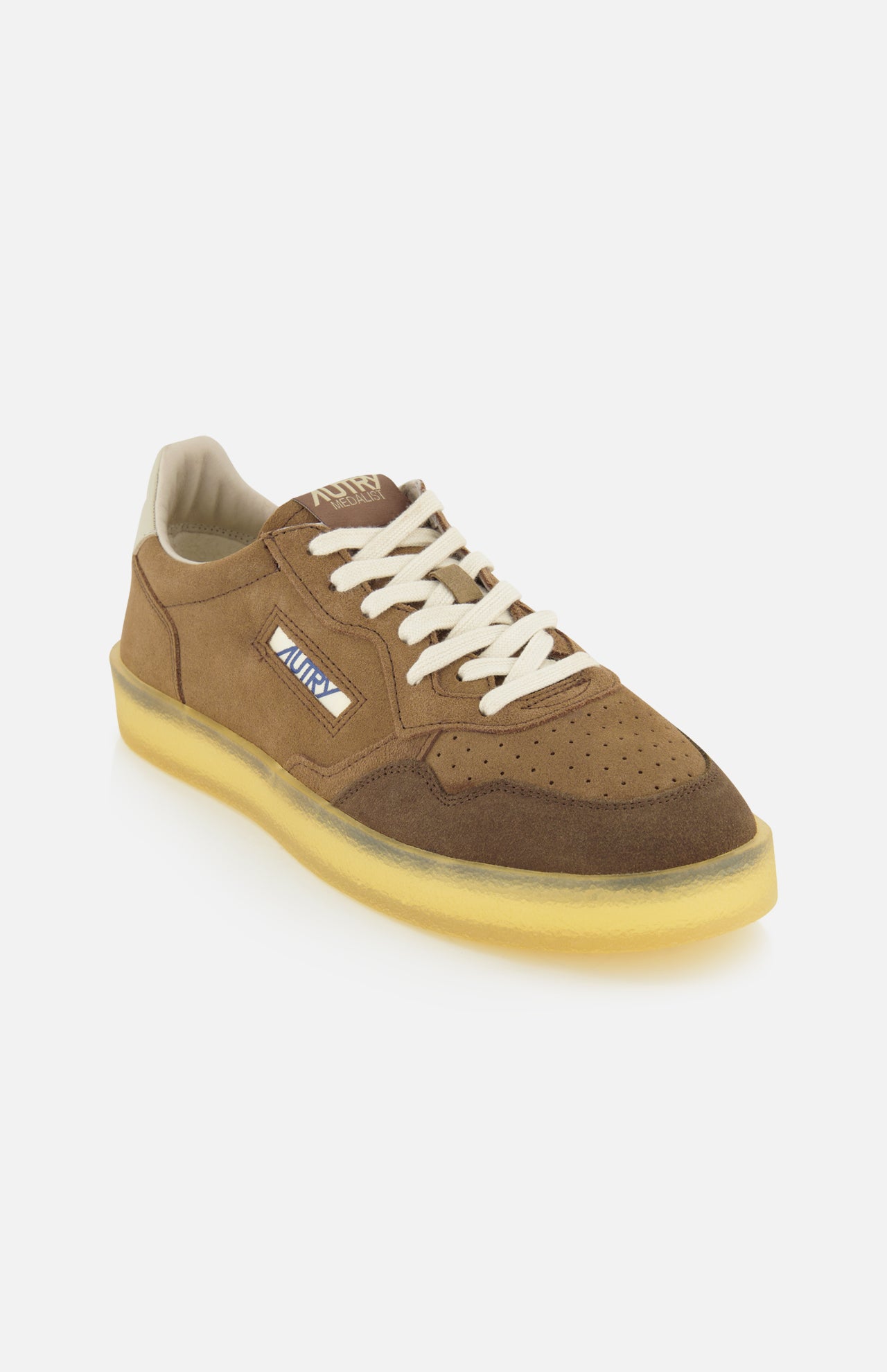 Introducing the AUTRY Medalist Low Man Suede Leather Sneaker—a classic brown suede low-top inspired by vintage tennis shoes. This stylish sneaker features white laces, a beige interior, and a translucent yellow sole. The side is adorned with the signature AUTRY logo patch and an American flag for an authentic touch. Angled slightly to the left, the shoe is showcased against a plain white background.