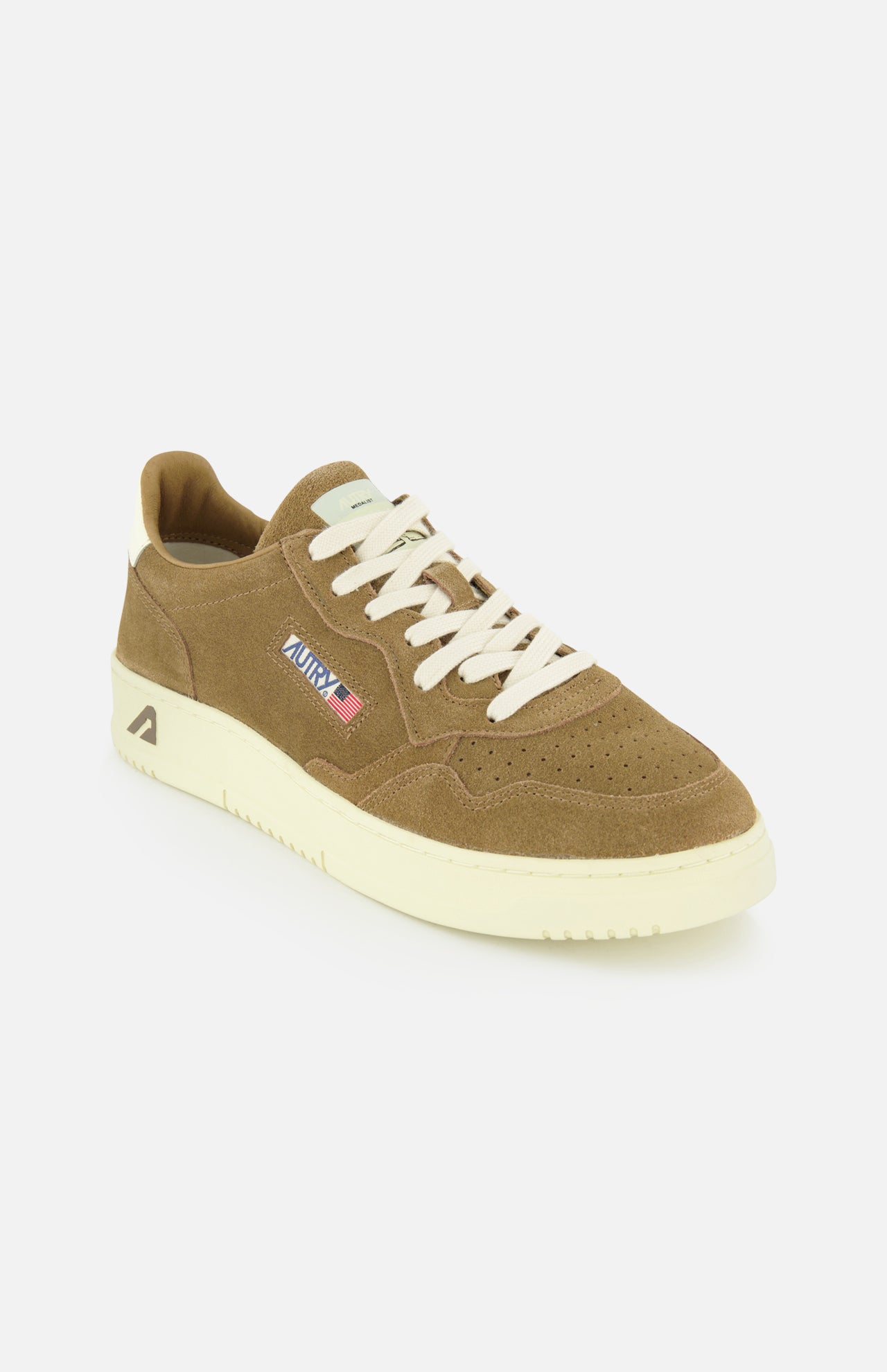 The AUTRY Medalist Low Suede Sneaker is a brown suede shoe inspired by 1980s tennis styles, featuring white laces, a thick off-white sole, and subtle embroidered flag and text details on the side, shown against a plain white background.