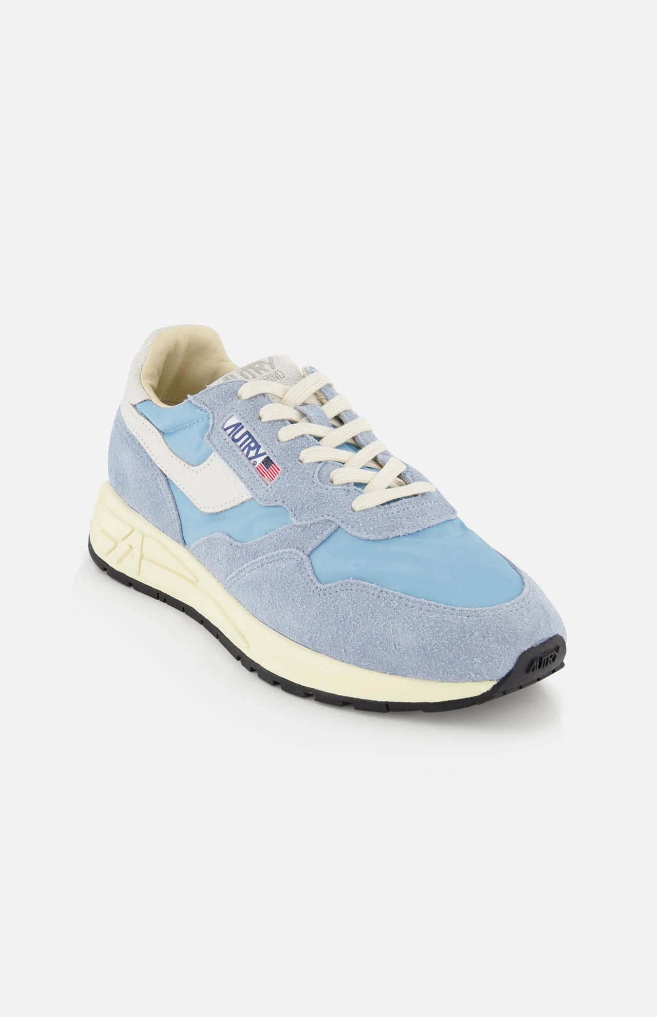 The AUTRY Reelwind Low Man Sneaker features a light blue and cream low-top design with white laces, a white and blue side logo patch, and a black sole—perfect for an active lifestyle—shown on a white background.