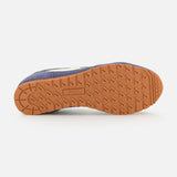 The image displays the sole of the AUTRY Windspin Low Suede Sneaker facing upward. Its light brown, textured grip is embossed with Autry, while the upper showcases a classic dark blue and white design inspired by 60s style.