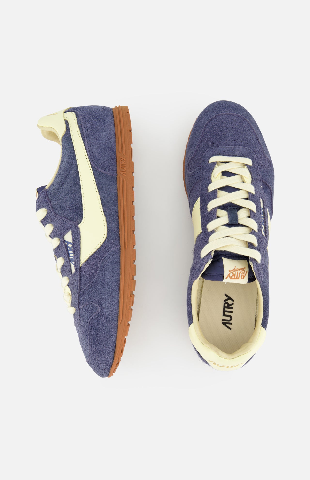 The AUTRY Windspin Low Suede Sneaker features blue suede uppers, off-white accents, and brown rubber soles. One 60s-inspired sneaker appears side-on; the other is shown from above against a plain white background.
