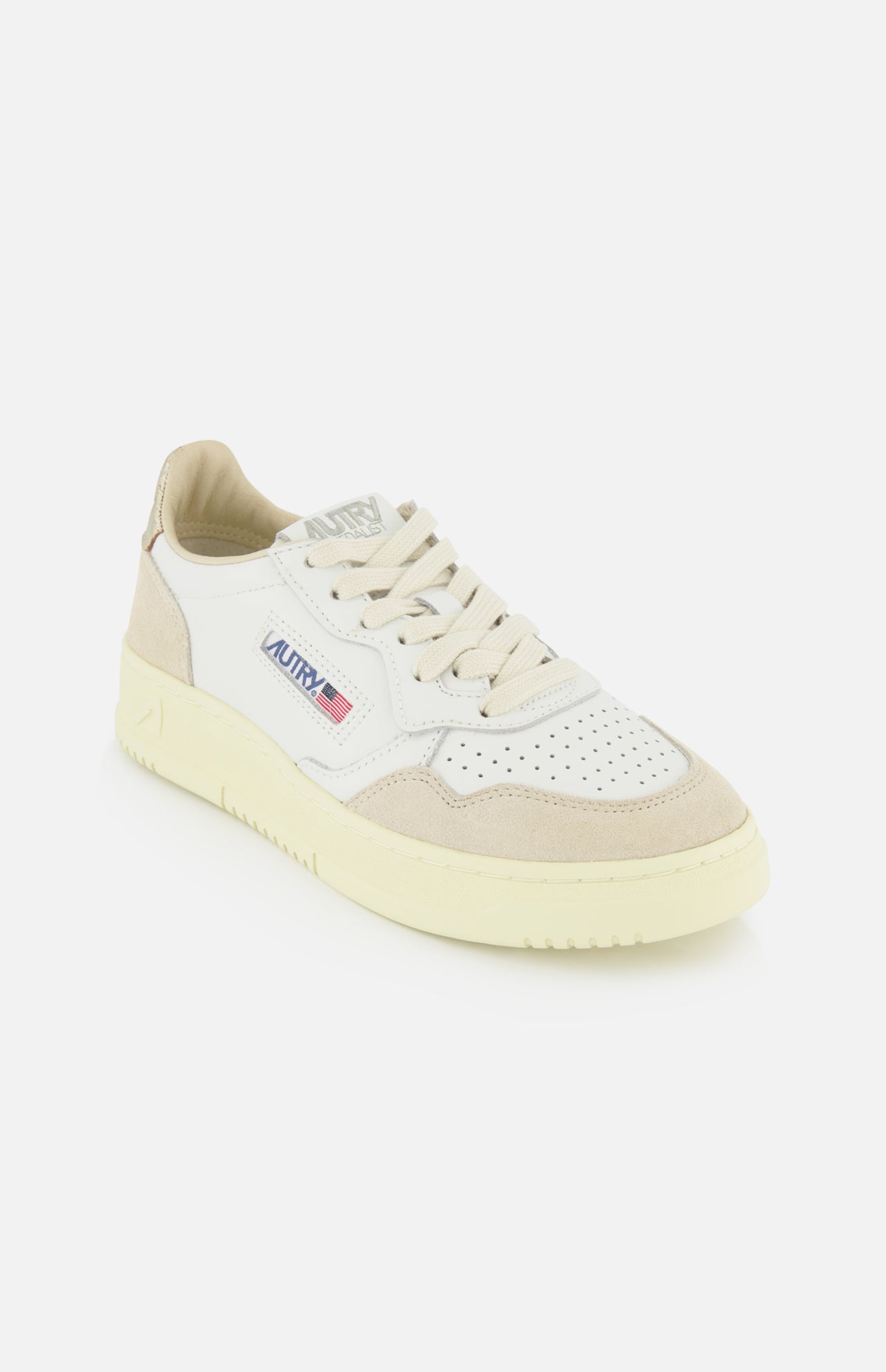 The AUTRY Medalist Low Gold Heel Sneaker features a white leather upper with beige accents, a perforated toe, cream platform sole, and the AUTRY logo with a small American flag on the side, capturing the vintage style of 1980s tennis shoes. It is displayed against a plain white background.