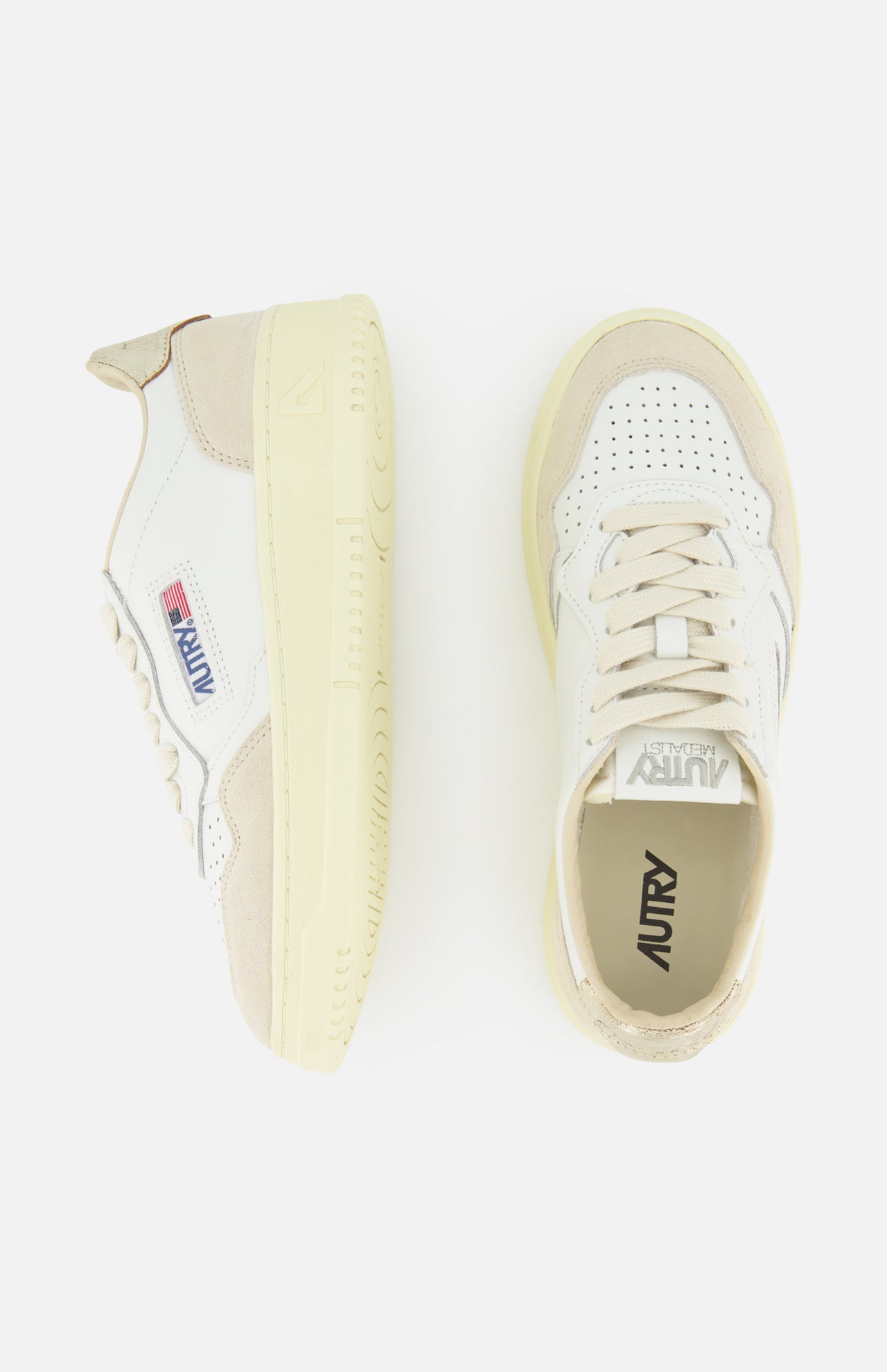 A pair of AUTRY Medalist Low Gold Heel Sneakers, shown from the side and top views, features suede accents, a perforated toe, and the signature American flag logo on the side. These stylish white leather sneakers with beige detailing are inspired by classic 1980s tennis shoes and are set against a crisp white background.