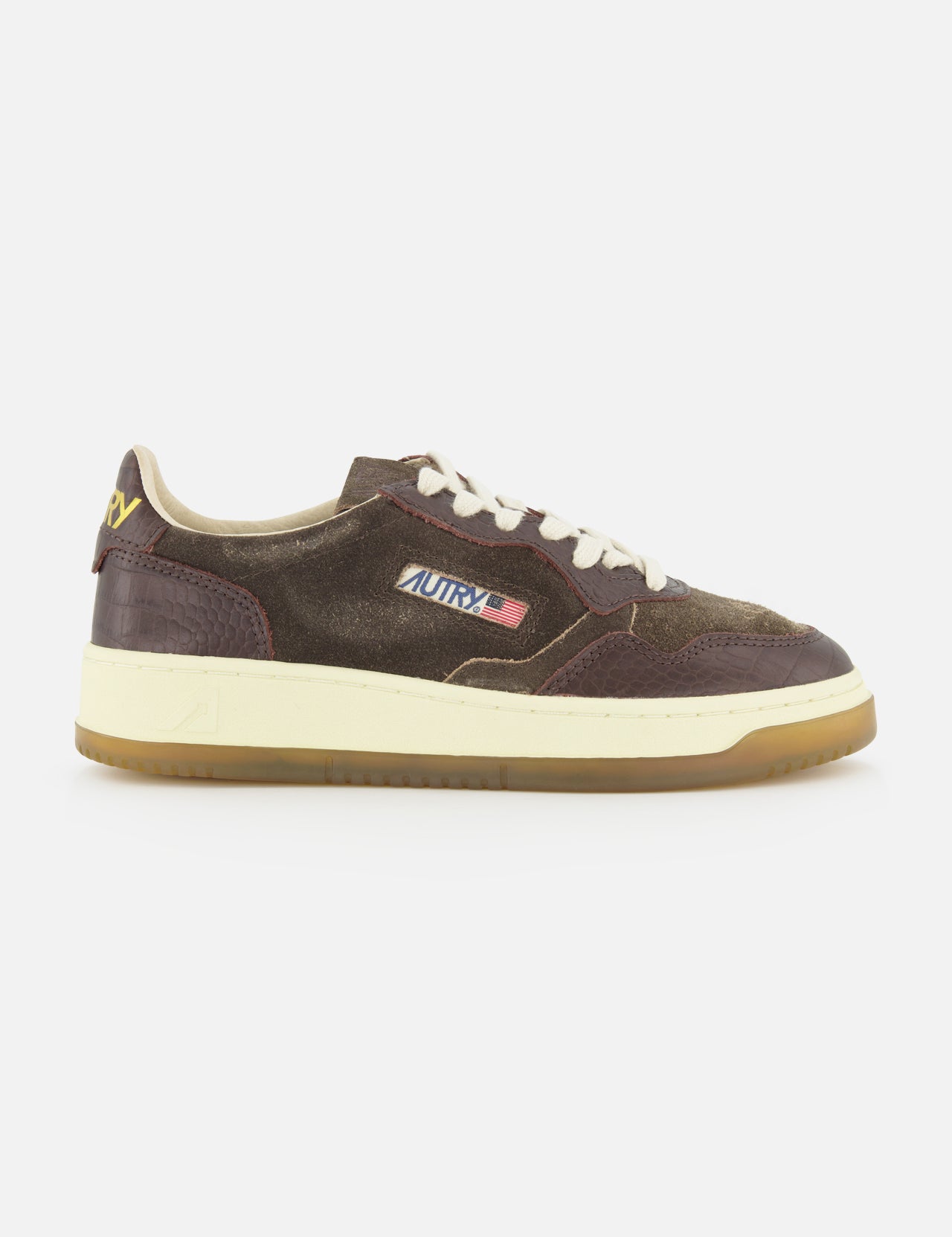 Introducing the AUTRY Medalist Low Suede Sneaker with Croco-effect: a low-top sneaker crafted from rich dark chocolate brown suede with distinctive croco-effect detailing. It features crisp white laces, a gum sole, and is finished with a small American flag logo on the side that reads AUTRY. Inspired by classic 1980s tennis shoes, this design effortlessly blends vintage appeal with contemporary style.